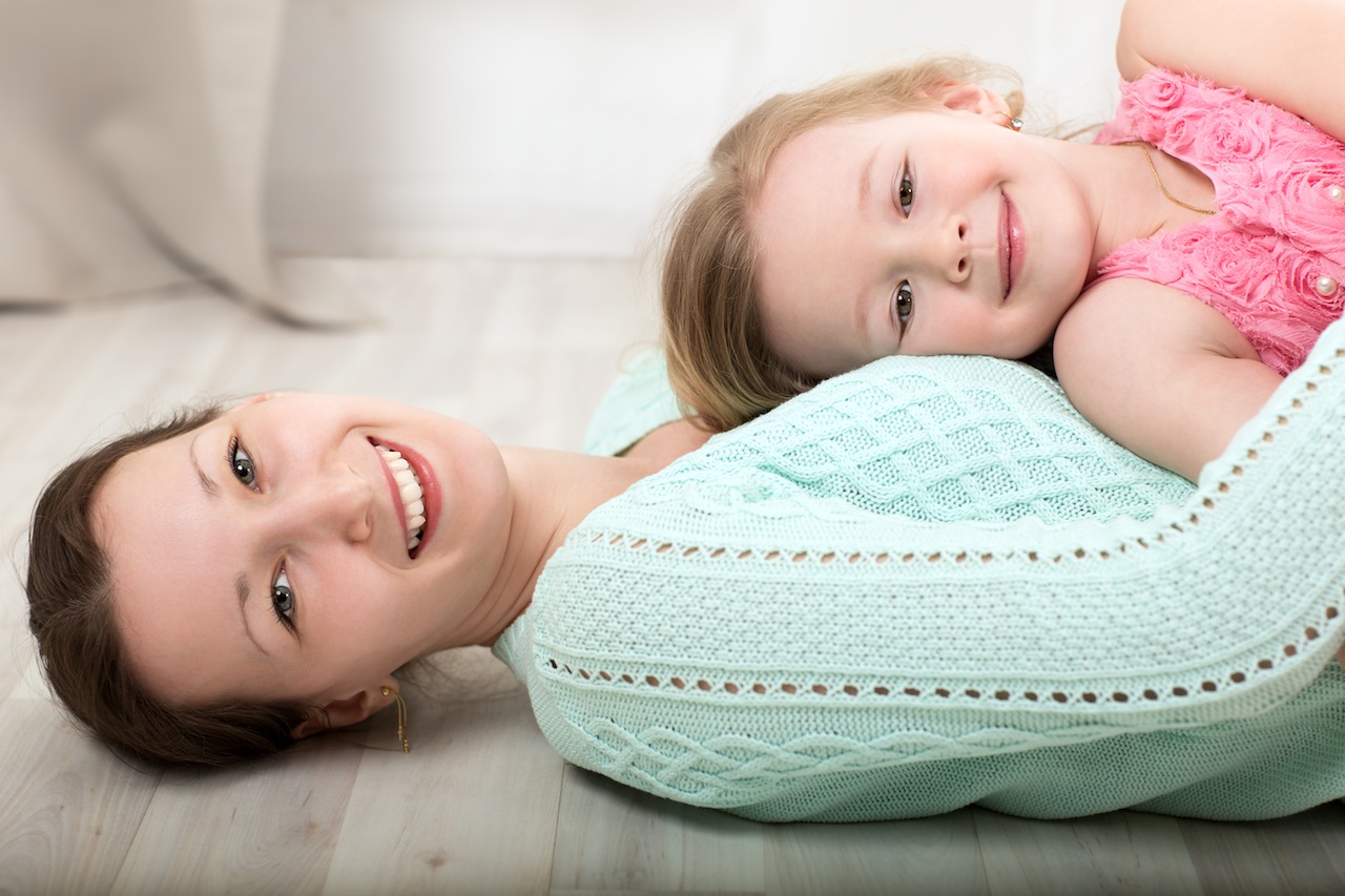 Happy young mother lying on the floor and holding smiling little daughter on the chest