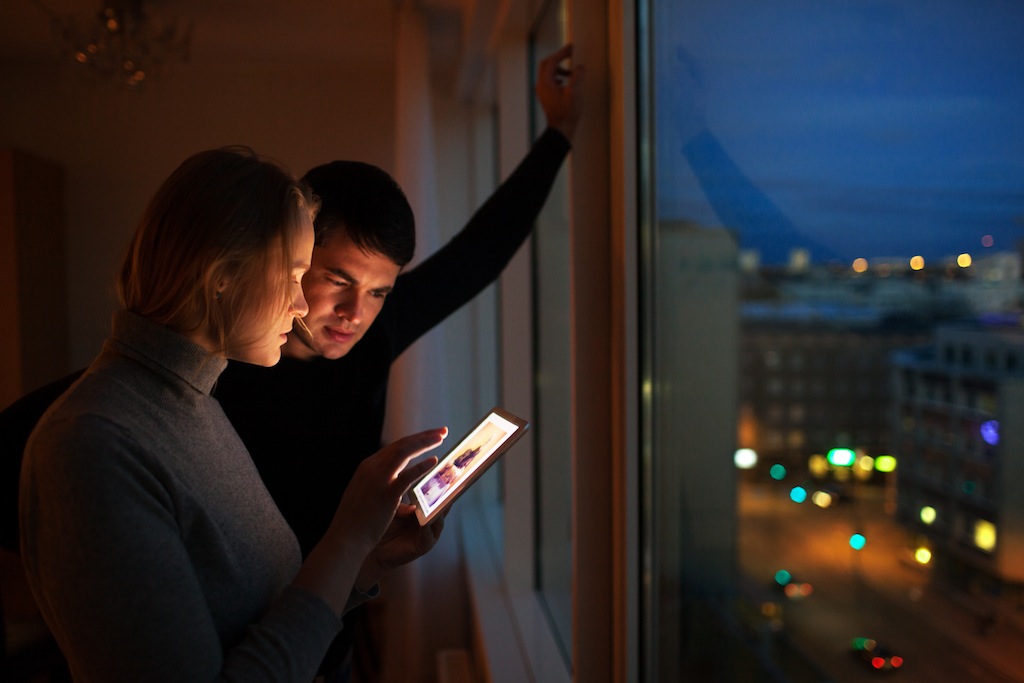 Young people using tablet computer at home. They looking at pictures standing by the window with the view of evening city