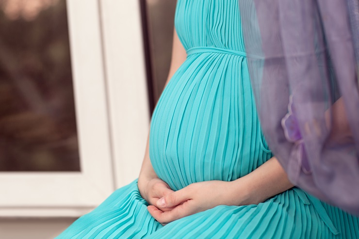 Close up of a pregnant woman wearing long blue dress sits on the window sill, face is unrecognizable